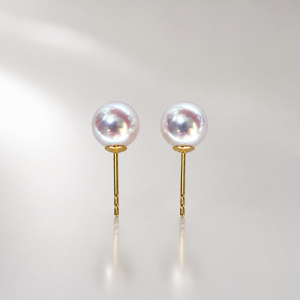 Pair of pale silver-white Akoya pearl stud earrings with gold-tone posts on a pearl-grey studio background